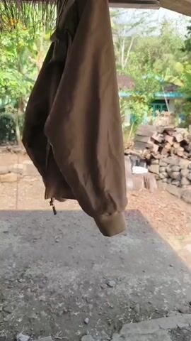 A brown jacket hangs from a clothesline. The jacket is slightly wrinkled and appears to be made of a thick material. The background shows a rural setting with trees, a house, and a pile of wood. The lighting suggests it is daytime.
