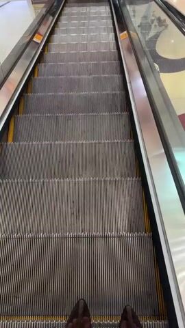 The video shows a first-person view of someone riding an escalator going down. The escalator is in a brightly lit shopping mall or similar commercial space. The escalator has metal steps with yellow safety strips along the sides. The railings are glass and metal. Other people can be seen on other floors of the mall in the background. The camera is focused downward towards the steps of the escalator.