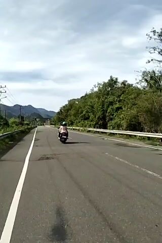The video depicts a motorcycle traveling on a rural road on a partly cloudy day. The road has a single white line marking the lanes, and a metal guardrail runs alongside it on the right. There are some potholes in the pavement. Lush, green vegetation covers the hillside to the right, contrasting with electrical poles and vegetation on the left. In the distance, mountain silhouettes can be seen beneath the sky. The motorcycle proceeds towards what appears to be a bridge or overpass.