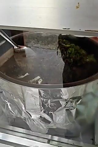 A close-up shot focuses on a food service setting. In the foreground, a metallic gray surface with some reflective quality is visible, partially covered with crumpled aluminum foil. Beyond this, a large round metal container holding a heap of noodles topped with shredded cabbage is seen, also wrapped in foil. To the left, another metallic surface and some debris are present. The background is largely obscured by a blurry, dark environment with some patches of light. The camera movement is subtle, suggesting a slow pan or slight shake.