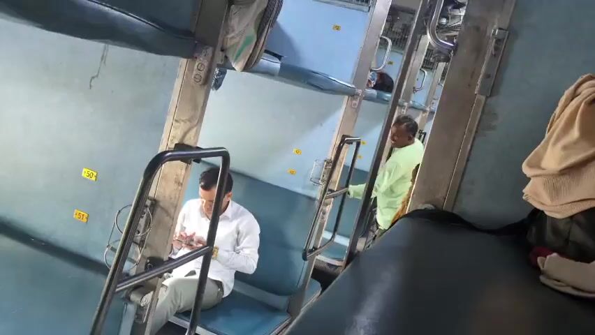 The video shows the interior of a train compartment with tiered sleeping berths. In the foreground, a person with dark curly hair and a red backpack is walking away from the camera. To their left, a man in a white shirt is seated on a blue berth, looking at his phone. Further back in the compartment, another man in a light green shirt is standing and looking towards the camera. A woman in a yellow sari is also visible standing near the man in green. The berths are stacked three high, and metal ladders are attached for access. The environment appears to be a travel setting, likely during the daytime given the lighting.