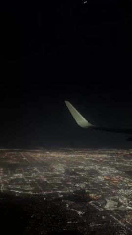 The video shows a view from an airplane window at night. The plane's wing is visible in the foreground, and below, a sprawling cityscape glitters with thousands of lights. The sky above is dark, with only a few visible stars or celestial objects.