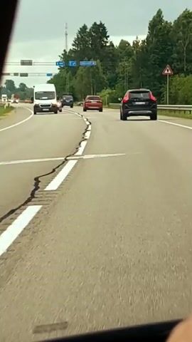 The video shows a first-person view from inside a vehicle driving on a highway. The road ahead has multiple lanes and is marked with white lines, some of which are cracked. Several vehicles are visible on the road, including a white van, a silver SUV, a red sedan, and a black SUV. Traffic appears to be moving at a moderate pace. Trees and vegetation line the sides of the road, and there are traffic signs and streetlights in the distance. The sky is overcast.
