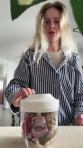 A woman with blonde hair stands in a kitchen and uses a white, hand-operated food chopper. She wears a black and white striped button-down shirt over a tan t-shirt. The chopper is clear and contains chopped vegetables, including red onions and tomatoes. The woman pulls the handle of the chopper repeatedly to further chop the vegetables inside. The background shows a modern kitchen with light-colored cabinets and a hanging light fixture.