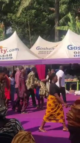 The video depicts a lively event, likely a celebration, taking place outdoors under white tents. People of diverse ages and attire, some dressed in traditional African patterns and colors, are seen dancing on a purple carpet. There are drummers in the crowd, contributing to the rhythmic atmosphere. The tents are branded with "Goshencity.com," along with phone numbers. The setting appears to be in a tropical or subtropical area, suggested by the lush green trees and palm-like foliage in the background.