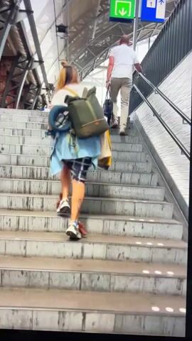 A woman with blonde highlights in her hair, wearing a white t-shirt, a blue jacket tied around her waist, and a plaid skirt over shorts, walks up a concrete staircase. She carries a green backpack and a blue neck pillow. Red laces are visible on her white sneakers. Another person, a man in a white shirt and khaki pants, walks ahead of her up the stairs. They are in what appears to be a train station, with signage and train tracks visible in the background.