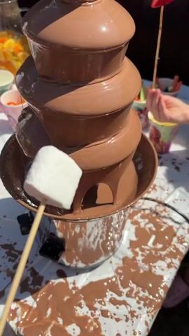 A close-up shot shows a chocolate fountain with marshmallows being dipped into it. The fountain is tiered, with chocolate cascading down from the top. Several marshmallows on skewers are visible, some partially submerged in the chocolate. In the background, there are cups and bowls of fruit and other dipping items, as well as children's hands reaching for them. The scene appears to be outdoors on a sunny day. Chocolate has spilled onto the white tablecloth covering the table.