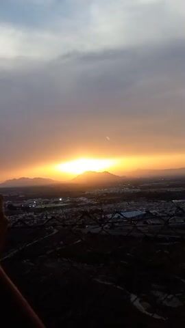 A sunset is captured from an elevated viewpoint, possibly a hill or building, looking out over a landscape of mountains and a town or city. The sun, a bright orange orb, is setting behind the mountains, casting a warm glow across the sky and illuminating the clouds. The foreground is dominated by a dark, blurry fence, suggesting the camera is positioned behind a barrier. Lights from the town or city are visible in the distance, twinkling like scattered diamonds. The sky transitions from a bright orange near the horizon to a darker, cloudy grey at the top of the frame. The overall impression is one of a serene and picturesque end to the day.