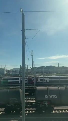 The video captures a bright, sunny daytime scene at a busy railway yard in Howald, Luxembourg, viewed from a moving train or vehicle.

In the foreground, several dark grey industrial tank cars are visible on parallel tracks. One prominent wagon features the white "VTG" logo and the partially visible text "radic," along with numerical identifiers such as "746 0 666-8." Hazard symbols, including a red diamond with a flame and a green diamond with a figure, are also present on the tank cars.

Further back, on adjacent tracks, multiple passenger trains are in view. One train is dark blue with bright green accents and displays "flou GrandEst" (partially obscured) on its side. Another passenger train, in a red and white livery, shows the "CFL" logo.

The railway infrastructure includes numerous overhead power lines (catenary system) and tall metal poles. Some poles are topped with bright lights, while others appear to be communication towers. In the background, beyond the railway lines, a collection of buildings with various architectural styles stands against a clear blue sky with minimal cloud cover. Distant trees are also visible.

An audible announcement, likely in French, can be heard in the background, mentioning "Bonjour" and "Luxembourg," indicating a railway station or train setting.