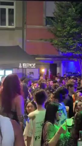 Here is a description of the video.

The video captures a lively outdoor gathering at night. A large crowd of people fills the area, with many faces visible, and they are dressed in casual clothing. The atmosphere is energetic, as people are dancing and enjoying themselves. In the background is a building, with a storefront that seems to be a bar or venue named "NOWV." The establishment has a black canopy set up in front, and there are colorful lights illuminating the facade. A tall tree stands to the right, adding some greenery to the urban scene.