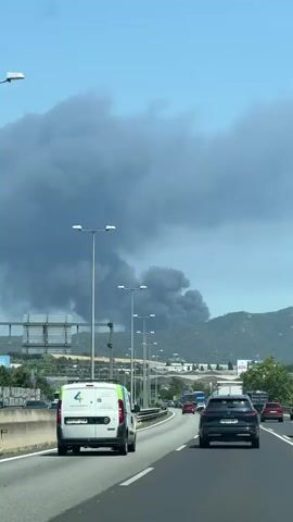 The video captures a daytime scene on a busy highway with multiple lanes filled with vehicles. In the distance, a large plume of dark grey smoke rises into the sky, partially obscuring the view of a mountain range. Streetlights line the side of the road, and various signs and buildings can be seen further away. The sky is partly cloudy, with patches of blue visible. The overall impression is that the video was taken from within a moving vehicle, capturing a concerning event in the distance.