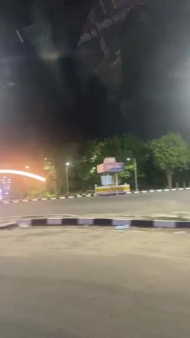 The video begins with a night view of a road intersection. A brightly lit archway is visible in the distance, adorned with lights. Various streetlights illuminate the area. To the right of the archway, a small structure with a blue and white sign is visible. A large billboard is prominently featured in the foreground, obscuring a portion of the scene. Trees line the sides of the road, and there are signs and poles on the left side. The camera pans to the right, showing a continuation of the road with more trees and streetlights. The lighting creates a vibrant and somewhat hazy atmosphere. The video ends with a blurred shot of trees and the sky. There are no people visible in the video.