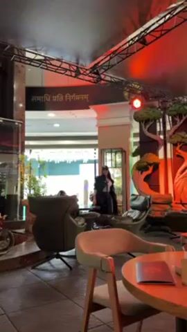 A woman is walking through a cafe. She is wearing a long black coat, a white top, black leather pants, and a baseball cap. The cafe has modern furniture, including round tables and comfortable chairs. There are decorative trees on the right side of the cafe. Above, there is a black metal structure with red lights. A banner in a foreign language is visible in the background.