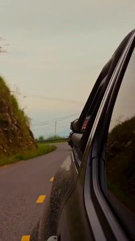 The video captures a scenic drive along a winding asphalt road in a rural, hilly environment, likely in Tân Quang Commune, Vietnam. The footage is shot from the perspective of a moving car, looking forward and slightly left. The road features yellow dashed lines.

On the left side of the road, a steep, grassy hillside is visible, adorned with lush green vegetation. The right side of the road is also lined with tall green grass and bushes, with a metal guardrail appearing in the distance. Overcast or hazy skies loom above, with distant hills or mountains framing the background. Power lines strung between wooden poles are a prominent feature of the landscape.

The main subject is a lone motorcyclist, wearing a white helmet and a bright red jacket, riding a touring or adventure-style motorcycle equipped with side luggage. The motorcyclist approaches from a left-hand curve, leaning into the turn as they navigate the road.

Notable details include a triangular warning sign on the right side of the road, commonly indicating caution or danger ahead. On the left, a white kilometer marker with red text displaying "H1 15" is visible as the motorcycle passes the camera. The overall lighting suggests an overcast day or possibly late afternoon.