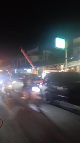 A blurry, night-time video shows a street scene with several vehicles. The camera appears to be in motion, possibly from a car. Headlights from oncoming traffic are bright and streaky. There are buildings lining the street, some with illuminated signs. A flag or banner, appearing red and white, is visible in the upper portion of the frame, attached to a pole and extending over the street. Vehicles, including a white van and a white car, are driving in the same direction as the camera. Another vehicle, possibly a motorcycle, with a bright headlight, is also visible in the foreground. The overall impression is of a busy street at night with moving traffic.