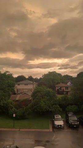 The video shows an outdoor view of a residential neighborhood under a heavily clouded, orange-tinted sky. In the foreground, a wet asphalt surface, likely a parking lot, is visible with two cars parked. One car is a silver sedan, possibly an Audi, and the other is a black SUV. Beyond the parking lot, there is a patch of green grass. Further in the background, several houses with pitched roofs are nestled among lush green trees. The sky is filled with dramatic, dark, and heavy clouds that create an eerie, atmospheric lighting condition, suggesting a storm or significant weather event is imminent or has recently occurred. The overall impression is one of a somber or intense weather situation.
