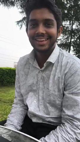The video shows a man from the chest up, wearing a light-colored shirt with a subtle pattern. He is outdoors, with greenery and trees visible in the background, suggesting a park or garden setting. The man is looking to his left and appears to be speaking, with his mouth open in some frames. The lighting suggests it may be overcast or late in the day. The man has dark hair and a beard. A hint of a laptop or tablet is visible at the bottom of the frame.
