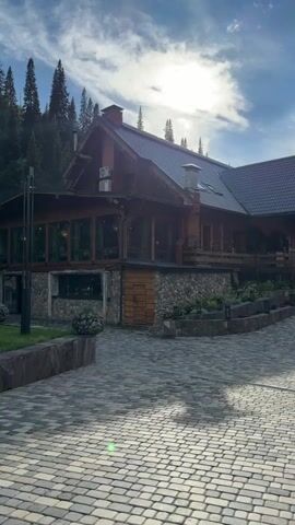 This video shows a large wooden and stone building with a pitched roof, nestled in a forest. The building appears to be a lodge or resort. A paved walkway leads up to the entrance. To the left of the building, there is a grassy area with two wooden swings and a hammock. A woman is visible on one of the swings, wearing a dark top and shorts. The background consists of a dense forest with tall evergreen trees and some deciduous trees. The sky is blue with scattered white clouds, and the sun is shining brightly. The overall impression is a serene and natural setting.
