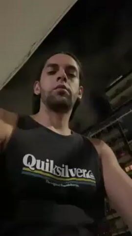 A man with long brown hair pulled back and a slight beard is outdoors at night. He is wearing a black Quiksilver tank top. He smiles slightly at the camera. He puts a cigarette to his mouth and takes a drag. A building and fencing are visible in the background.