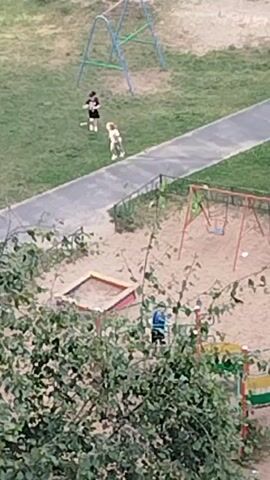 The video shows an aerial view of a playground. There are two children on a paved path. One child is wearing a black shirt with white stripes and white shorts, and the other is wearing a white shirt and light-colored shorts. They appear to be running. A sandbox is visible in the foreground, along with a swing set. Another child, wearing a blue shirt and black pants, is in the sandbox area. Trees and grass surround the playground.