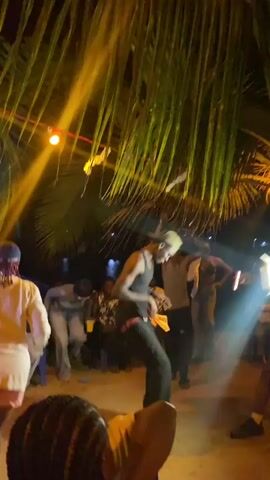 A nighttime beach party is underway, lit by spotlights and string lights hanging from palm trees.  A man in a tank top and baseball cap dances energetically in the sand in the foreground while others cheer him on, some recording with phones.  Some people are seated in the background.  A body of water is visible in the distance.