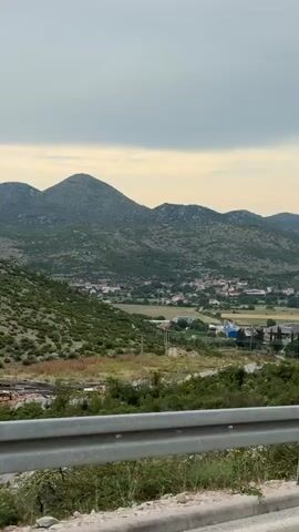 A video begins with a wide shot of a rural landscape. The foreground features a metal guardrail, partially obscured by green vegetation. In the middle ground, a small village with red-roofed buildings is nestled in a valley. Beyond the village, rolling hills covered in greenery rise towards the horizon. The sky above is overcast with muted gray clouds. The camera appears to be moving, suggesting it is capturing footage from a vehicle. No people are visible.