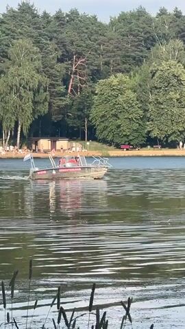 A metal boat with a canopy travels across a body of water. The boat has the words "СПАСАТЕЛЬНАЯ" and "ОСВОД" written on its side, indicating it is a rescue vessel. Two people wearing orange life jackets are visible inside the boat. The boat is moving from left to right in the frame. The background shows a treeline with various types of trees, and in the distance, there are buildings and some people on the shore. The water has ripples, suggesting the boat is creating a wake. The lighting indicates it is daytime, likely late afternoon or early evening due to the soft light.