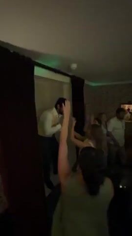 The video shows a lively indoor party. In the foreground, a person in a light-colored shirt and vest stands on an elevated platform holding a microphone, appearing to be singing or addressing the crowd. Several people are visible in the background, some dancing with their hands raised. The environment is dimly lit, with colorful lights projected onto the ceiling, including red, blue, and green patterns. A bar area is also visible, with bottles and glasses on the counter, and DJ equipment in the foreground. The overall atmosphere suggests a celebration, possibly a wedding reception or a party.