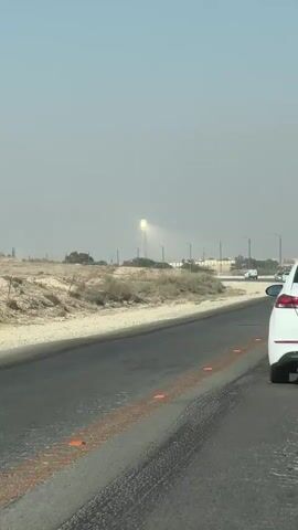 The video shows a view from inside a vehicle traveling on a two-lane road. The road is paved and has yellow and orange markings. The landscape is dry and appears to be a desert or arid region with sparse vegetation and sand. In the distance, a tall tower with a bright light emanating from its top is visible, illuminating the hazy sky. Several cars are driving on the road in the same direction as the camera vehicle. The weather appears to be sunny with some haze in the air.