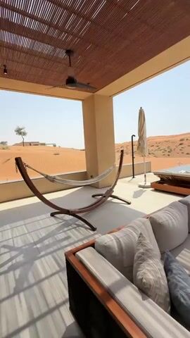 This video shows a high-angle exterior shot of a luxurious outdoor living space overlooking a desert landscape. The scene features a comfortable, modern sofa with plush cushions in the foreground. To the left, a wooden hammock stand with a striped hammock is visible. Beyond the seating area, there is a swimming pool with lounge chairs and an umbrella. The background consists of vast sand dunes under a clear blue sky. The architecture includes a wooden slatted roof providing shade over the patio area, with a ceiling fan attached. The overall impression is one of serene and upscale desert relaxation.