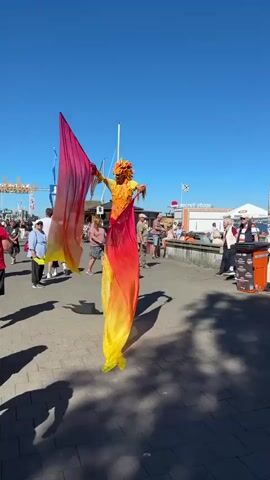 A person on stilts, dressed in a colorful costume resembling a phoenix with fiery orange and yellow fabric, is walking along a paved walkway. The stilt-walker holds large, flowing fabric streamers in shades of pink, orange, and yellow, which billow in the wind. They have a mask that appears to be part of the phoenix costume.

The scene is set outdoors on a sunny day with a clear blue sky, suggesting it is daytime. A crowd of people is visible in the background, strolling along the walkway and sitting at tables. Some are dressed casually for warm weather. There are tents and structures, one with a sign that partially reads "secret stage."

Further in the background, there are boats docked or moored, visible on the water, and more buildings along the waterfront. The overall atmosphere appears to be festive, likely a public event or festival taking place near a waterfront. The paved ground is made of interlocking bricks, and shadows from people and the stilt-walker are cast on it. The location context indicates this is Kiel, Germany.