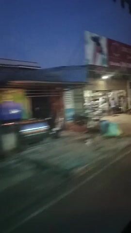 The video shows a street scene at dusk or dawn. The camera appears to be moving, possibly from inside a vehicle, as evidenced by the motion blur. Along the side of the road, there are several storefronts. One store is open, revealing shelves stocked with various items, possibly clothing or merchandise. A few people can be seen inside the open store. Further down the street, other buildings are visible, some with their shutters down. The sky is a deep blue, suggesting it is either early morning or late evening. There is street lighting visible, illuminating the road and the storefronts. The overall impression is of a quiet, ordinary street in a commercial area as the day is ending or beginning.