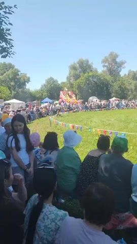 A crowd of people are gathered in an outdoor grassy area on a sunny day, watching an event. In the foreground, several people are seated or standing, facing away from the camera. Behind them, a group of people are visible on the grass, some of whom are standing and watching. Further back, there is a line of tents and booths, and beyond that, a large crowd is seated on the grass, facing a central area. In the distance, a person is running across the grass, and a flagpole is visible. There is a bouncy castle in the background. A string of colorful triangular flags stretches across the grassy area. The overall atmosphere appears festive.