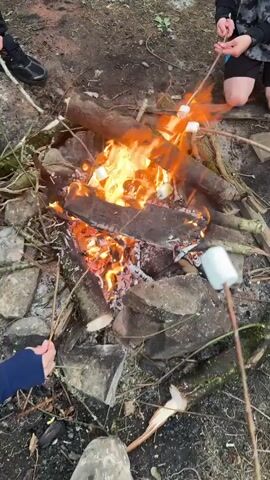 This video shows a close-up of a bonfire with several marshmallows being roasted over the flames. Three people are visible in the frame, though only their lower extremities are shown. One person is wearing a black shoe, another dark shorts, and the third, white sneakers. The setting appears to be outdoors, likely a campsite or a wooded area, with dirt, rocks, and fallen branches forming the ground. The fire is a central element, with logs and embers glowing brightly and flames leaping upwards. The marshmallows, skewered on sticks, are positioned near the fire, with some beginning to brown and char. The lighting suggests it could be late afternoon or early evening, as there is no harsh direct sunlight, and the fire provides a warm glow. The overall mood is casual and relaxed, indicative of a typical outdoor activity like camping or a backyard gathering. There is no visible text in the video. The location context provided is Lettergunnell, Ireland.