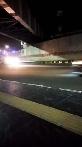 A vehicle with bright headlights drives under an overpass at night. The road is wet, reflecting the headlights. A sidewalk is visible in the foreground with tactile paving. The overpass has a concrete structure with a pattern of circular indentations.