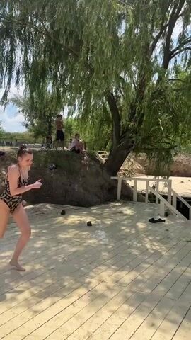 A young woman in a black and gold patterned swimsuit runs and leaps from a wooden dock into a body of water. She is captured mid-air, with her arms outstretched and body crouched as she prepares to enter the water. Another person is already in the water, swimming towards the splash zone created by the woman's dive. Further to the right, a man with sunglasses is swimming and looking towards the divers. The scene takes place on a sunny day with a blue sky dotted with white clouds. The water is clear and green, and the surrounding area features a rocky embankment with sparse vegetation. A large willow tree with drooping branches provides shade over the wooden dock in the foreground. In the background, other people can be seen relaxing on the grassy bank. The overall atmosphere appears to be one of recreation and enjoyment on a warm day. There is no visible text in the media file.