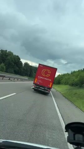 A car driving on a highway in overcast conditions films a red truck ahead in the same lane. The truck has "Constantin" written on the side with a "C" logo above. A gray car passes the truck on the left. There is vegetation on both sides of the highway with guardrails on the left. A blue sign indicates "Fuchsberg" with a gas pump symbol. The video is shot from the driver's perspective.