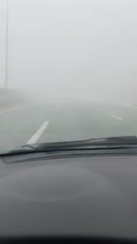 The video depicts a view from inside a car driving on a highway blanketed in thick fog. The dashboard is visible in the foreground. The road ahead is barely visible through the heavy fog, with faint white lines marking the lanes. Windshield wipers are present but not in use. The overall environment suggests poor visibility conditions due to the dense fog.