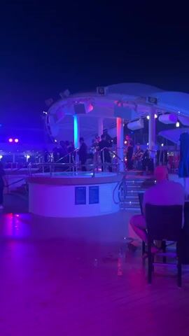 The video shows a band performing on a circular outdoor stage at night. The stage is illuminated with blue and pink lights. Several musicians are visible on the stage, playing instruments such as a guitar and drums. A few people are dancing on the stage. In the foreground, a person is sitting on a chair, facing away from the camera. The ground in front of the stage appears to be wet, with reflections of the lights visible on the surface. The sky is dark, indicating it is nighttime. The overall atmosphere appears to be festive and lively.