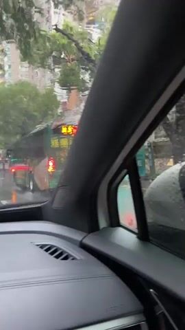 The video, filmed from inside a car, shows a street scene during rainfall. In the foreground, a side mirror is visible with raindrops on its surface. Through the side window, a green bus is passing by, with some characters visible on its destination board. The view then shifts to the front windshield, revealing a street lined with trees and buildings. Several cars are on the wet road, their headlights and taillights creating reflections on the wet pavement. The overall atmosphere is cloudy and wet, indicating continuous rain.