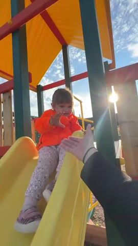 A young child wearing an orange hooded sweatshirt and patterned leggings is sitting on a yellow plastic slide on a playground. The child is looking towards the camera and holding out a hand, which is met by an adult's hand reaching into the frame. The playground has a yellow and red structure with green supports. The ground is covered in gravel and mulch. In the background, houses and trees are visible under a blue sky with some clouds. The video captures the child in different moments on the slide and then walking on the playground.