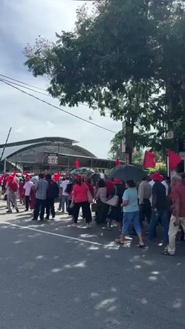 A daytime video clip from Delkanda, Sri Lanka, shows a large group of people marching down a street. Many participants are wearing red shirts or caps and carrying red flags, suggesting a protest or political rally. They walk in the same direction, occupying a significant portion of the road.

The street is lined with buildings, including multi-story structures that appear to house businesses. Visible signs on these buildings include "Castrol," "BOBACULT Bubble Tea," and partially "INDOVINDI MOTORS" and "BLACK." Lush green trees with dense foliage are also visible along the roadside, providing shade. Power lines crisscross above the street.

The weather appears clear and sunny, with a bright sky and some scattered clouds. A white SUV is parked on the side of the road as the group passes. The scene depicts an organized public gathering in an urban environment.