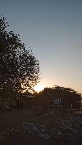 The video shows a sunset scene along a body of water. On the left side, there is a tree with a dense canopy and a small structure with a thatched roof. Below the tree, there are several bicycles leaning against something. In front of the structure, there are people gathered. Further to the right, there are more structures on stilts over the water, with a wooden pier extending into the water. There are several boats moored near the pier. The water is calm with gentle waves. The sky is a gradient of light blue at the top, transitioning to a soft orange and pink hue near the horizon where the sun is setting. The foreground on the left appears to be a dark sandy or rocky area with debris. The overall atmosphere suggests a tranquil evening by the sea or a large lake.