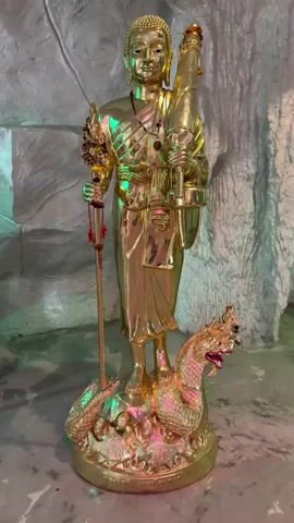 The video opens with a static shot of a golden statue of a figure standing on a mythical creature. The statue is adorned with robes, a necklace, and holds a staff in its left hand and a vessel in its right. The background is a rough, rock-like textured wall illuminated with green and pink lights, creating an ambient, cave-like atmosphere. A gong hangs on a wooden stand to the right of the statue, also lit by the colored lights. The camera then pans slightly to the right, showing more of the gong and the illuminated background. The scene shifts to the right further, revealing a person sitting in a rattan chair with a fan in front of them. The person is wearing a hat and appears to be observing something off-camera. The colored lighting continues to dominate the scene, casting shadows and highlights. The video appears to be a brief tour or showcase of a dimly lit, decorated space with religious or cultural significance.