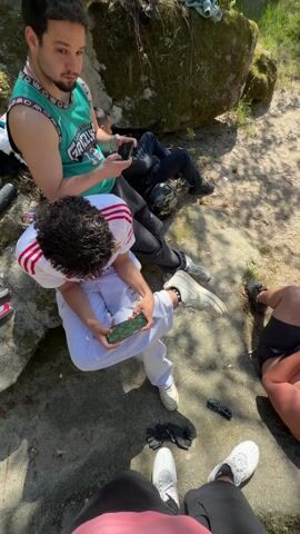 The video shows a group of young men enjoying a sunny day outdoors. They are sitting on a rocky ground area near a wooded area with a view of a river. Some of the men are using their phones, and one man is wearing an aluminum foil hat. The camera then pans to show a view of the river with lush green trees and vegetation surrounding it. Then the camera returns to the group of men, focusing again on the man with the foil hat who is still using his phone.
