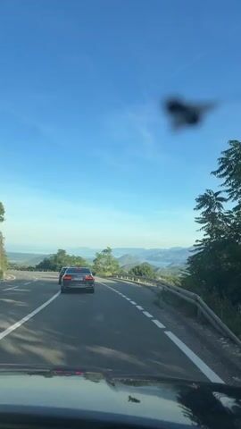 The video shows a view from inside a vehicle traveling on a paved road. The road curves to the left and descends slightly. To the left of the road, there is a steep embankment with green vegetation and scattered trees. To the right, there is a metal guardrail and a scenic overlook with a view of mountains and water in the distance. The sky is clear and blue. A dark-colored sedan is visible ahead, driving in the same direction. The dashboard of the vehicle from which the video is recorded is partially visible in the foreground.