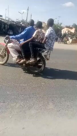 Three men are riding on a motorcycle. The man in the front is driving. The man in the middle is sitting behind the driver. The third man is sitting behind the second man. They are on a street with buildings and shops on the side. The sky is clear and the sun is shining. There is a police car on the side of the road.