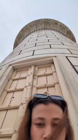 The video shows a low-angle shot of a person's face in the foreground, with a tall, white, cylindrical structure behind them. The structure appears to be made of white stone blocks with dark lines separating them, and it has a decorative top. There is a large, cream-colored door or panel on the structure, with a grid pattern of lines and metal hardware. The person in the foreground has dark hair and is wearing sunglasses on top of their head. The sky is visible in the background and appears overcast. The camera is held close to the person's face, creating a somewhat distorted perspective. The video is very short, lasting only a few seconds, and there is no discernible action or interaction taking place.
