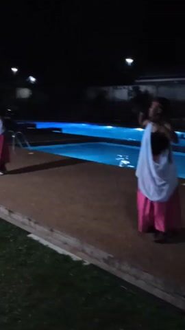 The video shows three people performing a traditional dance by a pool at night. The dancers are wearing dark tops, pink skirts, and white shawls draped over their shoulders. They are moving in a coordinated fashion, their arms and bodies flowing with the music. The pool is lit with a bright blue light, and the surrounding area is dark except for a few streetlights. The dancers are on a stone patio next to the pool, with a grassy area behind them. The audio includes the sound of drumming, singing, and cheering, adding to the festive atmosphere.
