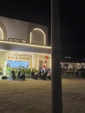 A nighttime view of a building facade with a sign that reads "HELLO BEACH COFFEE & RESTO". The building has arched entrances and is lit by decorative string lights. Several motorcycles are parked in front of the building. People are walking in the foreground and near the entrances of the building. A red and white striped flag is visible on a flagpole to the right of the building. The ground in front of the building appears to be dirt or sand.